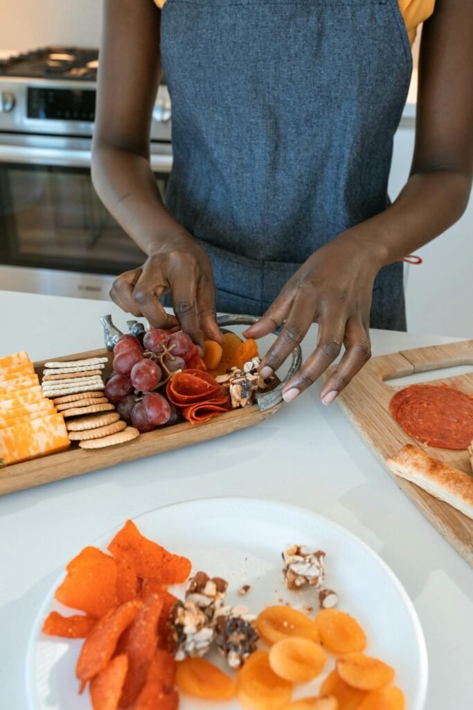 Person arranging a variety of cheeses, meats, and fruits on a charcuterie board indoors.
